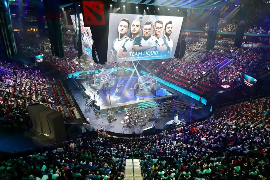 Image of Dota 2 tournament with people attending and each team on the monitors.